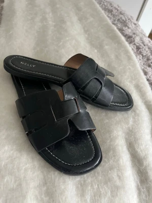 Svarta skor/sandaler från Nelly - Stilrena svarta slip-in sandaler från Nelly med breda flätade remmar och fyrkantig tå. Tillverkade i konstläder med låg sula och vita sömmar runt kanten. Perfekta för en avslappnad och trendig look. Parfekta sommar skor