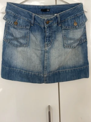 Blå jeanskjol från H&M - Säljer en klassisk blå jeanskjol från H&M med två stora fickor fram och två bak. Kjolen har knappstängning och markerade sömmar för en cool denimlook. Perfekt till sneakers eller boots för en avslappnad stil.