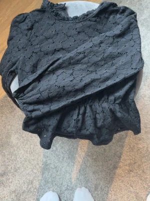 Svart spetsblus med volang - Snygg svart blus i spets med volang nertill och diskret mönster. Blusen har långa ärmar och en liten volang vid halsringningen. Perfekt för dig som gillar en romantisk och trendig stil.