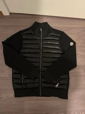 Svart cardiganjacka från Moncler - Snygg svart cardiganjacka från Moncler med quiltad framsida i blankt material och stickade ärmar och rygg. Jackan har dragkedja framtill, två fickor med dragkedja och en Moncler-logga på ärmen. Perfekt för dig som gillar stilren och modern design.