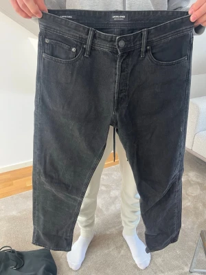 Svarta jeans från Jack & Jones - Säljer ett par svarta jeans från Jack & Jones med klassisk femficksmodell och raka ben. Jeansen har knappgylf och bälteshällor, tillverkade i denim med en lätt tvättad look. Perfekta för en avslappnad och stilren outfit.