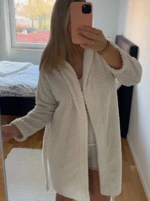 Vit fluffig morgonrock - Mysig vit morgonrock i fluffigt och mjukt material med struktur. Modellen har långa ärmar, sjalkrage och knytskärp i midjan. Perfekt för att chilla hemma och hålla sig varm under kyliga morgnar.