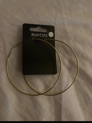 Stora guldfärgade hoops Montini - Säljer ett par stora, tunna hoops i guldfärg från Montini. Örhängena är cirkelformade och har enkel, stilren design som verkligen syns. Perfekta för dig som gillar statement-smycken och vill addera lite extra till din look.