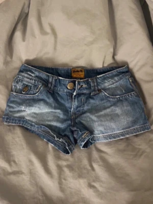 Blå jeansshorts från Sicka 79 - Säljer ett par klassiska blå jeansshorts från Sicka 79, knappgylf och snygga slitningar. Shortsen har uppvikta benslut och dekorativa sömmar på bakfickorna. Perfekta för varma dagar och enkel att matcha med allt.