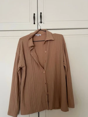 Beige ribbad kofta  - Säljer en beige ribbad kofta med knappar framtill och krage. Koftan har långa ärmar och är tillverkad i ett mjukt, stretchigt material som känns skönt mot huden. Perfekt för lager på lager och enkel att matcha med olika outfits. Oversized