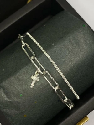 Silverarmband med kors och pärlor - Två stilrena armband i silverfärg, ett med större länkar och ett hängande kors dekorerat med små pärlor, samt ett smalare armband täckt av gnistrande pärlor.