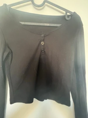 Svart croppad topp med knappar - Snygg svart croppad topp med lång ärm och tre knappar framtill. Toppen har en enkel och stilren design med lätt v-ringad halsringning. Perfekt att matcha med höga jeans eller kjol.