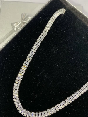 Glittrigt tennis-halsband i silver - Ett elegant tennis-halsband med rektangulära, gnistrande stenar i rad. Halsbandet har en klassisk och stilren design i silverfärg, perfekt för att lyfta vilken outfit som helst. 