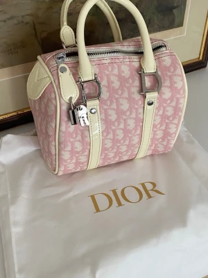 Rosa Dior monogram handväska - Säljer en söt rosa handväska från Dior med klassiskt monogrammönster och krämvita detaljer i läder. Väskan har två handtag, silverfärgade metalldetaljer, dragkedja och hänglås med nyckel. Perfekt för dig som vill ha en ikonisk och trendig accessoar. Säljer den relativt billigt eftersom jag inte har några äktenhetsbevis och kan därav inte garantera äktenhet. 