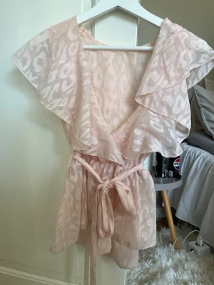 Ljusrosa omlottopp med volang - Söt ljusrosa omlottopp med volangdetaljer och knytband i midjan. Toppen har ett subtilt mönster, är ärmlös och har en luftig, flowy passform. Perfekt för dig som gillar romantisk och feminin stil.