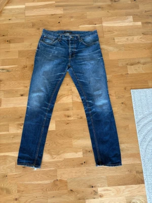 Blå jeans med slitningar och raka ben - Säljer ett par blå jeans med slitningar och raka ben. Jeansen har klassisk femficksdesign, ljusa detaljer och råa kanter vid benslut. Materialet är denim och färgen är mörkblå med ljusare partier. Perfekt för dig som gillar en avslappnad och trendig look.