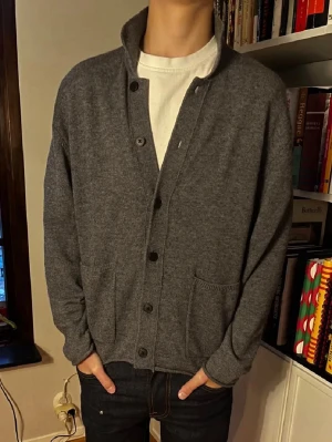 Mörkgrå Massimo Dutti kofta med knappar - Mysig mörkgrå kofta från Massimo Dutti med knappar framtill och två stora fickor. Koftan har hög krage och är tillverkad i ett mjukt stickat material. Perfekt att slänga på över en t-shirt för en avslappnad look. Har en liten defekt med lite målarfärg vid insida handled.