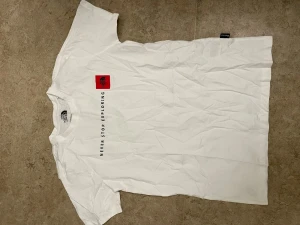 Vit The North Face t-shirt - Säljer en vit t-shirt från The North Face med trycket 'Never Stop Exploring' i svart text och en röd logga på bröstet. Klassisk passform och rund hals. Perfekt för dig som gillar enkel och stilren design.