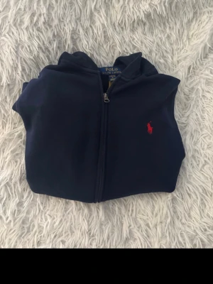 Mörkblå hoodie från Polo Ralph Lauren - Mörkblå hoodie med dragkedja från Polo Ralph Lauren. Klassisk design med huva, känguruficka och röd broderad logga på bröstet. Mjuk bomullskvalitet som är skön att ha på sig.