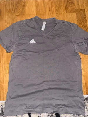Grå v-ringad t-shirt från Adidas - Snygg grå t-shirt från Adidas med v-ringning och korta ärmar. Diskret Adidas-logga på bröstet. Tillverkad i mjuk bomull som känns skön mot huden. Perfekt till jeans eller shorts för en avslappnad look.