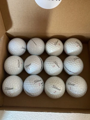 Titleist pro v1x A/B klass  - Säljer ett set med 12 vita golfbollar från Titleist, med klassisk dimple-design och märkning i svart och rött. Några bollar har siffror och en har en liten smiley. Perfekt för golfentusiasten som vill ha kvalitetsbollar.