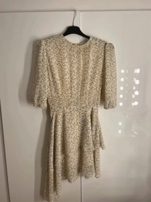 Blommig kortklänning med puffärm - Supersöt kortklänning i ljusgul med små gula blommor och diskreta gröna detaljer. Klänningen har puffiga trekvartsärmar och en lätt utsvängd kjo som är asymmetrisk . Perfekt för sommarens alla tillfällen! Materialet känns lätt och luftigt.