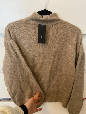 Beige stickad tröja från Another Story - HELT NY. Mysig beige stickad tröja från vero Moda.med hög krage och långa ärmar. Tröjan har en enkel, stilren design och är ribbstickad upptill. Perfekt för lager på lager och en avslappnad look. Jättelen 