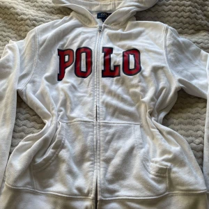 Vit hoodie från Ralph Lauren - Säljer en vit hoodie från Ralph Lauren med stor POLO-text i rött och svart på framsidan. Tröjan har dragkedja och långa ärmar, perfekt för en avslappnad stil. Den är tillverkad i ett mjukt material och har en klassisk passform. Nån liten fläck som säkert går bort. Storlek L i barn som passar för xs. Skriv gärna innan köp😊
