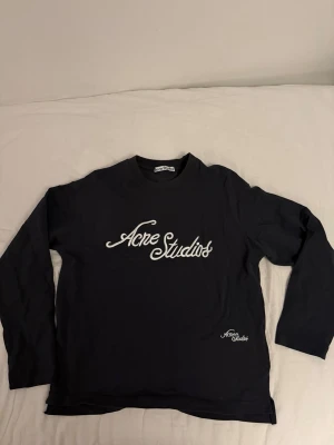 Grå långärmad tröja från  Acne Studios - Snygg grå långärmad topp från Acne Studios med vit logotyp framtill och mindre logga vid nederkanten. Klassisk rund halsringning och relaxed fit. Perfekt för dig som gillar stilrena och trendiga plagg. Ser blå ut på bilden men är mer grå i verkligheten, endast testad. Hör av er vid frågor!