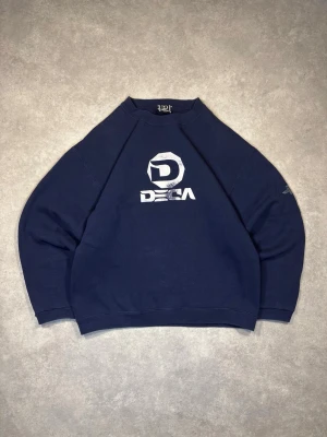 Vintage DECA sweatshirt - Snygg mörkblå sweatshirt från DECA med tryckt logga i vitt på bröstet. Tröjan har rund halsringning, långa ärmar och ribbade muddar vid ärmslut och nederkant. Perfekt för en avslappnad och cool stil. Materialet känns mjukt och bekvämt.