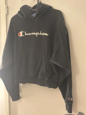 Svart hoodie från Champion - Svart hoodie från Champion med broderad logga på bröstet och ärmen. Klassisk känguruficka framtill, ribbade muddar och huva med snörning. Perfekt för en avslappnad och sportig stil.
