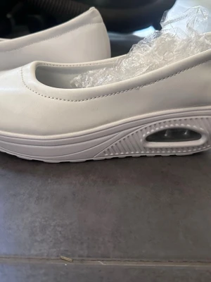 Vita ballerinaskor med platåsula - Säljer ett par vita ballerinaskor med modern platåsula och luftbubbla i sulan. Skorna har rund tå och är tillverkade i ett slätt syntetmaterial. Perfekta för dig som vill ha en sportig och trendig look.