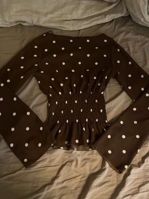 Brun prickig blus Gina Tricot XS - Supersnygg brun blus från Gina Tricot med vita prickar och smockad midja. Blusen har långa, vida ärmar och en lätt stretchig känsla i materialet. Perfekt för dig som gillar trendiga detaljer och en feminin siluett.