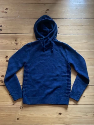 Kashmir hoodie - Mörkblå stickad hoodie med huva och dragsko. Tröjan har ribbade muddar vid ärmslut och nederkant samt ett diskret stickat mönster längs sidorna. Perfekt för dig som gillar en enkel men snygg look.