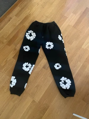 DENIM TEARS BLACK - Säljer ett par svarta mjukisbyxor med stora vita blommönster över hela byxorna. Byxorna har resår i midjan och vid bensluten för en skön och avslappnad passform. Perfekta för chill dagar eller när du vill sticka ut lite extra.