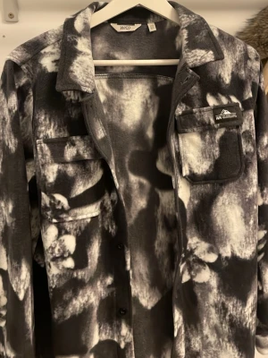 Svartvit overshirt från WND - Overshirt i mjukt material från WND med svartvit batikmönster. Skjortan har två bröstfickor med lock, knäppning framtill och klassisk krage. Perfekt för lager på lager och ger en cool streetwear-look.