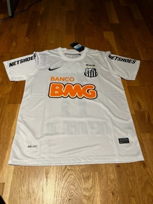 Santos 11/12 Neymar jr #11 - Vit fotbollströja från Nike för Santos FC med Neymar Jr och nummer 11 på ryggen. Tröjan har svarta och orange tryck, klubbmärke på bröstet och flera sponsorer. Tillverkad i lätt och ventilerande Dri-FIT-material, perfekt för träning eller match.