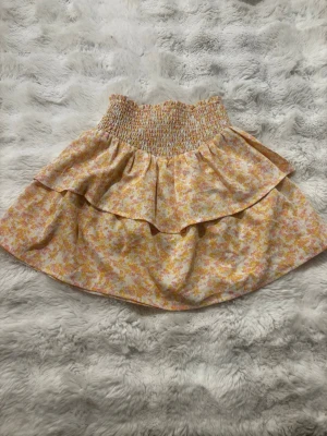 Blommig volangkjol med smock - Supersöt kort kjol med blommigt mönster i ljusa toner av orange, rosa, gul och vit. Kjolen har bred smock i midjan och dubbla volanger för en extra flowy känsla. Perfekt för dig som gillar en feminin och trendig stil.