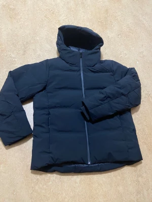 Svart pufferjacka med huva Uniqlo  - Säljer en svart pufferjacka med hög krage och stor huva. Jackan har en rak passform, långa ärmar och dragkedja framtill. Perfekt för kalla dagar när du vill hålla dig varm och stilsäker.