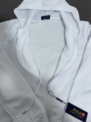 Vit hoodie från Polo Ralph Lauren - Snygg vit hoodie från Polo Ralph Lauren med dragkedja framtill och klassisk huva med dragsko. Ikoniska loggan broderad på bröstet. Tillverkad i mjukt material som känns skönt mot huden. Perfekt för en clean och stilren look. Byxor följer med också, helt ny dress