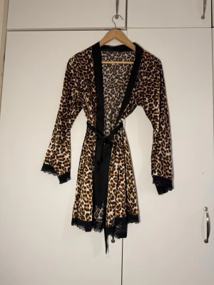 Leopardmönstrad morgonrock med spets - Snygg morgonrock med leopardmönster och svarta spetsdetaljer vid ärmslut och nederkant. Svart kant och knytskärp i midjan. Perfekt för en chill dag hemma eller när du vill känna dig extra lyxig. ( bandet kan ej fästas vid rocken men går att knyta)