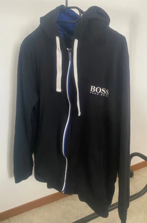 Svart hoodie från Hugo Boss - Svart hoodie med dragkedja från Hugo Boss. Stora vita snören i huvan och vit logga på bröstet. Klassisk passform med långa ärmar och mjukt material. Perfekt för en chill och stilren look.