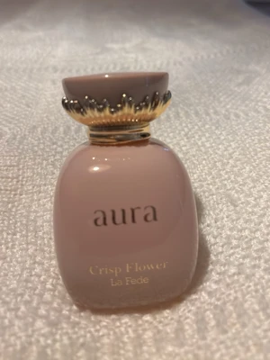 Aura Crisp Flower parfymflaska - Säljer en elegant parfym från La Fede med namnet Aura Crisp Flower. Flaskan är ljusrosa med gulddetaljer och har en unik, dekorativ kork som ger en lyxig känsla. Perfekt för dig som gillar blommiga dofter och snygg design på parfymhyllan.