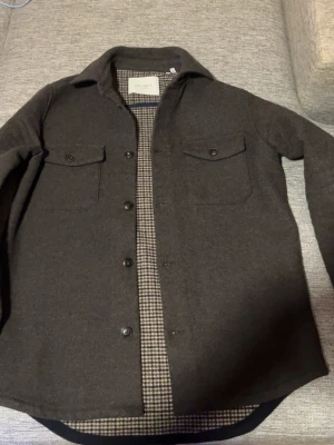 Mörkgrå overshirt från Les Deux - Säljer en snygg mörkbrun overshirt från Les Deux med klassisk krage och två bröstfickor med lock. Insidan har ett stilrent hundtandsmönster. Perfekt att bära som lagerplagg. Materialet känns tjockt och varmt, troligen ullblandning.