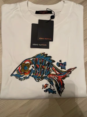 Vit Louis Vuitton t-shirt med fiskmotiv - Säljer en vit t-shirt från Louis Vuitton med ett färgstarkt fiskmotiv på framsidan och ett tryck med Louis Vuitton-loggan i blått och rött på baksidan. T-shirten har rund hals och är kortärmad. Materialet är mjuk bomull och designen är riktigt unik.