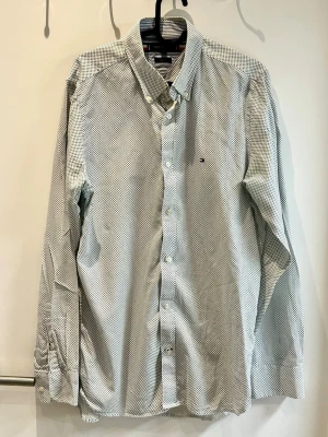 Vit prickig skjorta från Tommy Hilfiger - Snygg vit skjorta från Tommy Hilfiger med små svarta prickar och klassisk knappstängning. Skjortan har lång ärm och en liten broderad logga på bröstet. Perfekt för dig som gillar stilrena detaljer och tidlös design.