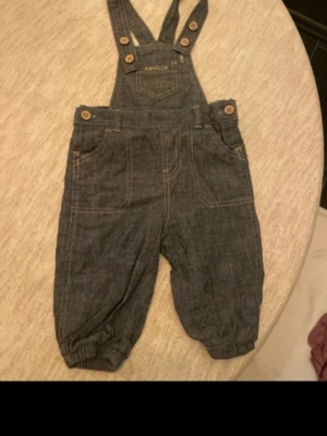 Mörkblå denim jumpsuit från Newbie - Supersöt mörkblå denim jumpsuit från Newbie med justerbara axelband och träknappar. Fickor fram och bak samt resår vid benslut för extra komfort. Perfekt för en trendig och avslappnad look.