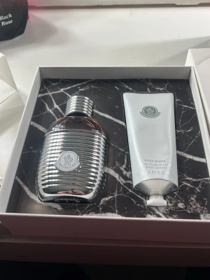 Moncler Pour Femme parfymset - Lyxigt parfymset från Moncler Pour Femme med en elegant parfymflaska i silverfärgad metall och tillhörande body cream i matchande tub. Snygg presentask med marmorerad insida och Moncler-logga. Perfekt för dig som gillar exklusiva dofter och stilren design.