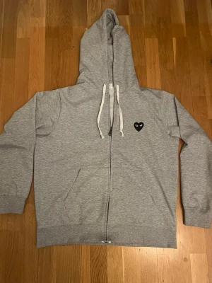 Comme des Garcons play hoodie - Grå zip-hoodie från Comme des Garçons Play med vit dragsko i huvan och fickor framtill. Liten svart hjärtlogga med ögon på bröstet och stor svart hjärtdetalj med ögon på ryggen. Storlek L men passar även M 