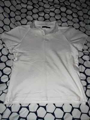 Vit piké t-shirt med krage - Klassisk vit piké t-shirt med krage och knappar framtill. T-shirten har korta ärmar och är tillverkad i ett mjukt bomullsmaterial. Perfekt för en stilren och enkel look.