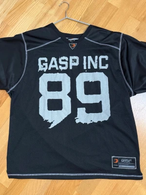  Mesh topp från GASP INC - Sportig svart mesh topp från GASP INC med stora siffror '89' och logga framtill. Oversized passform och korta ärmar, perfekt för en streetwear-look. Materialet är luftig polyester och sömmarna är markerade i kontrastfärg.