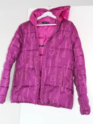 Lila pufferjacka från Champion - Snygg lila pufferjacka från Champion med heltryckt logotyp i rosa över hela jackan. Jackan har en vadderad design, dragkedja framtill och en matchande huva med blank finish på insidan. Perfekt för kalla vinterdagar och riktigt trendig look.