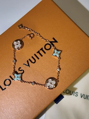Louis Vuitton armband i roséguld - Snyggt armband från Louis Vuitton i roséguld med ikoniska LV-loggor och blommotiv. Armbandet har detaljer i ljusblått och glittrande stenar, samt kedja med små pärlliknande inslag. Perfekt accessoar för att lyfta din stil.With box💝