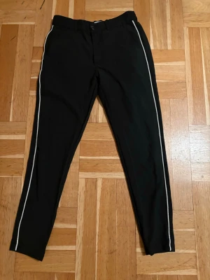 Svarta mjukisbyxor med vita ränder - Svarta mjukisbyxor med vita ränder längs sidorna. Dragkedja och knapp framtill. Fickor både fram och bak. Endast använda 2-3 ggr.