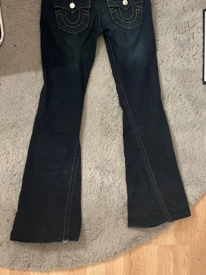 Mörkblå bootcut jeans med kontrastsömmar - Säljer ett par mörkblå bootcut jeans med snygga kontrastsömmar och dekorativa fickor bak med vita knappar. Jeansen har klassisk femficksdesign och utsvängda ben, perfekta för en avslappnad men trendig look.